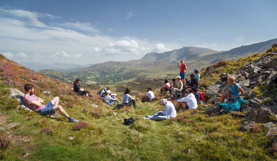 Group of hikers on mental health retreat seated on grass in welsh hills for mindfulness 