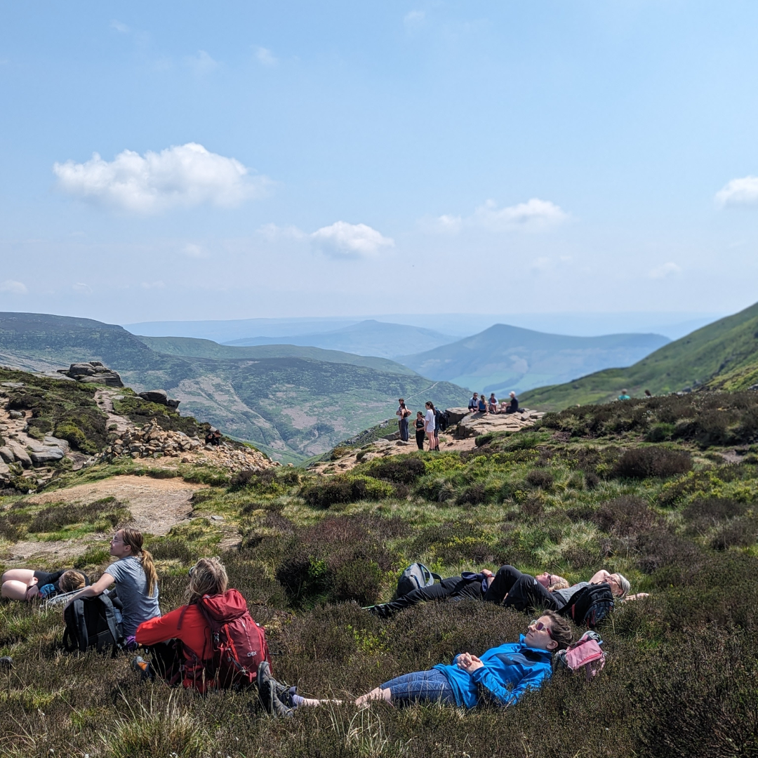 People relaxing on a grassy hilltop with a scenic view of mountains under a clear blue sky.