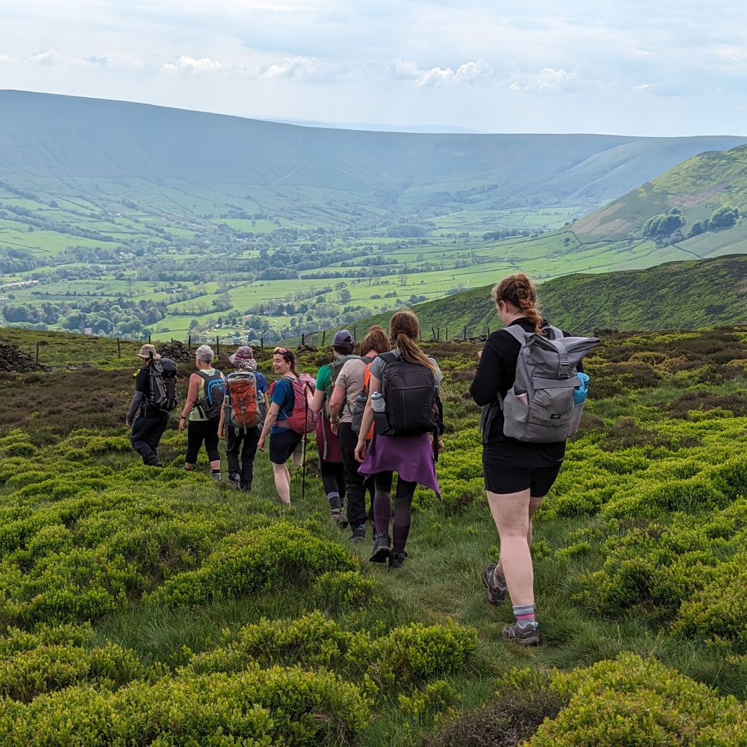 A group of hikers with backpacks walking along a path through green heather and grass on a mountain ridge, with a valley and hills stretching into the distance under a cloudy sky.