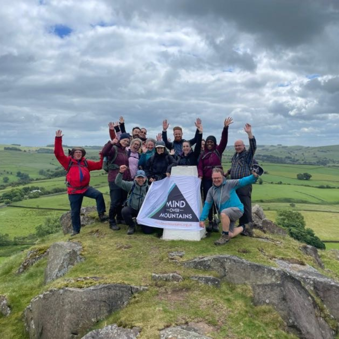 A group of hikers posing together on a grassy rocky summit holding a white 'MIND OVER MOUNTAINS' banner, with green fields and a cloudy sky in the background.
