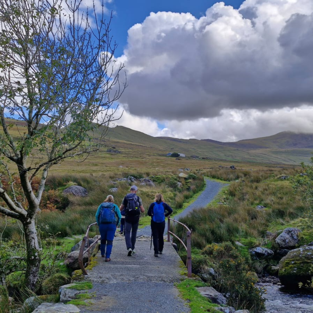 Three hikers walk across a small stone bridge on a path through a grassy mountain landscape under a blue sky with large white clouds.
