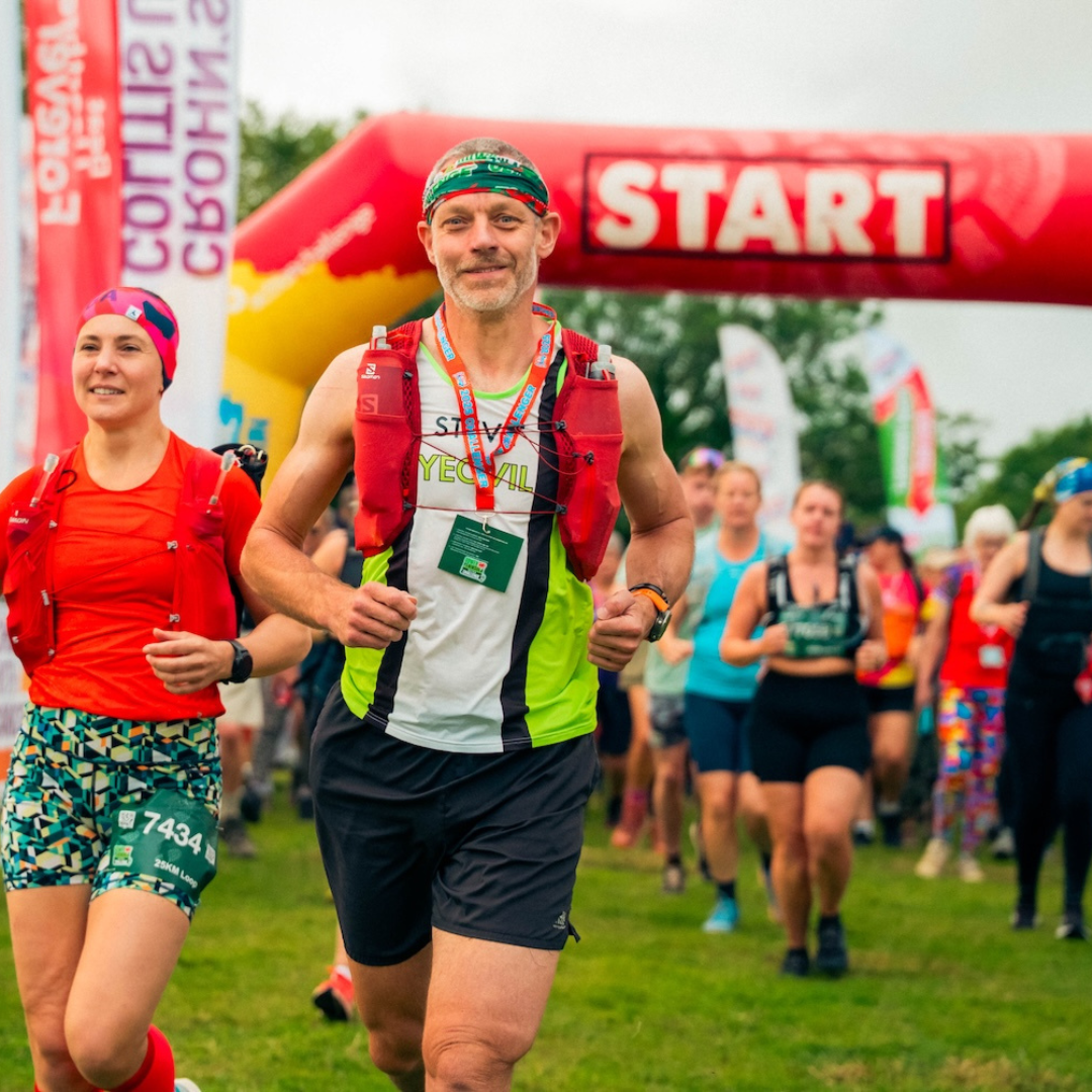A male runner in a bright neon green and white top, black shorts, and a red hydration pack crosses a red 'START' arch at a race, with other competitors and grassy background visible.