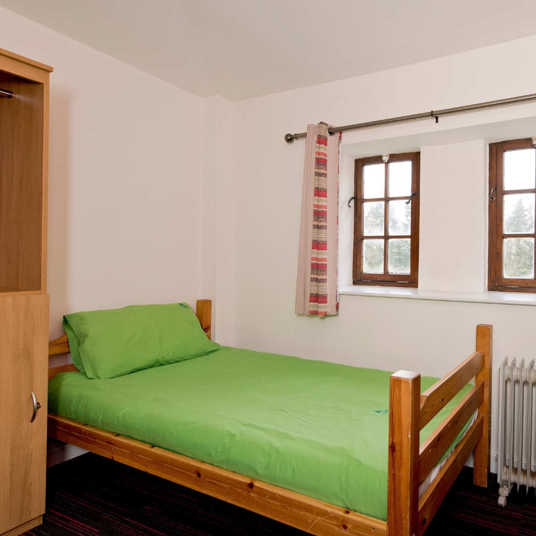 Bedroom with a wooden bed and green bedding, light-colored walls, and a window with curtains.
