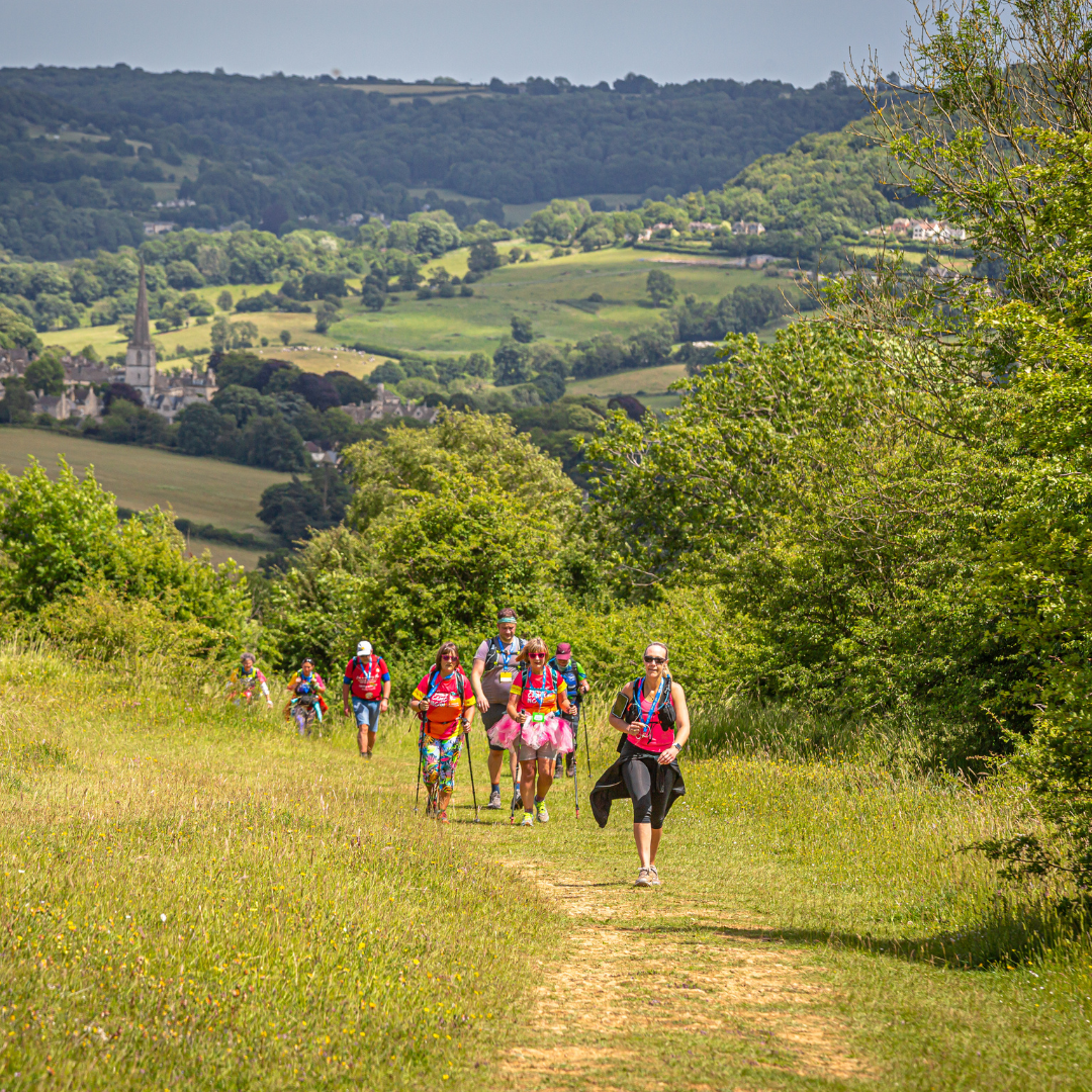 A group of trail runners or hikers walks along a dirt path in a lush green valley with rolling hills and trees in the background.