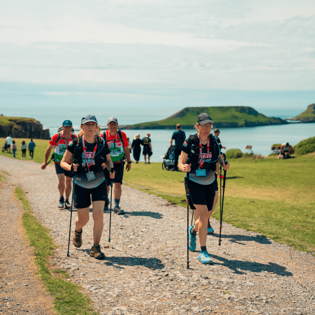 Two trail runners wearing bibs and hydration packs walk along a gravel path with ocean and green hills in the background.