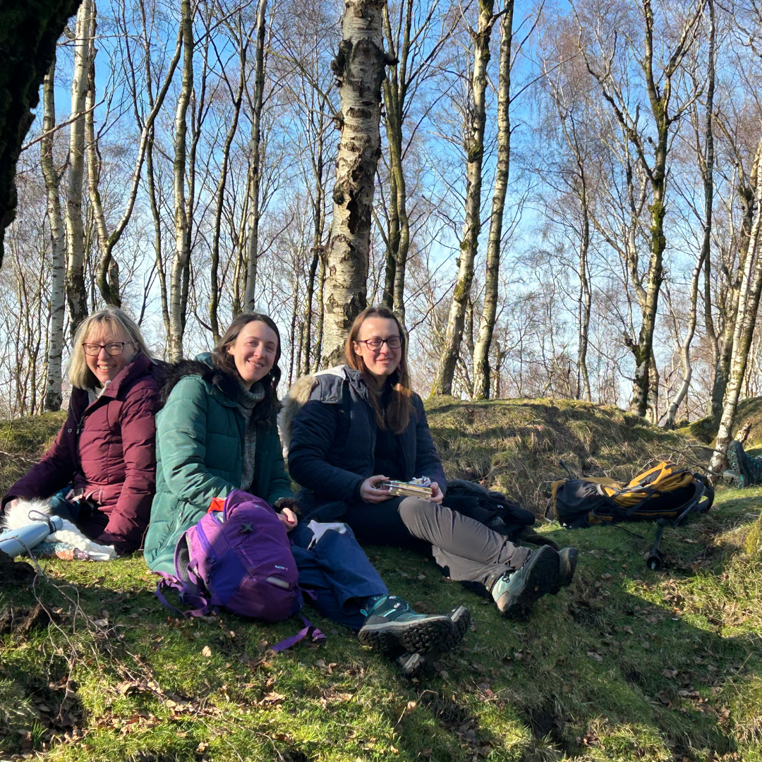 Three women sitting on a grassy hill in a birch forest, with backpacks beside them in a sunny outdoor setting.