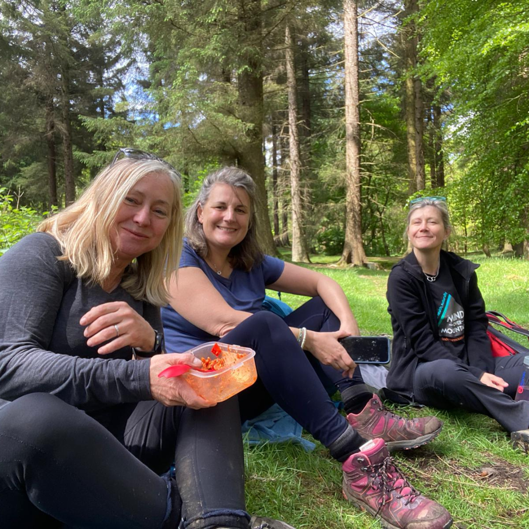 Three women sitting on the grass in a forest, enjoying a snack.
