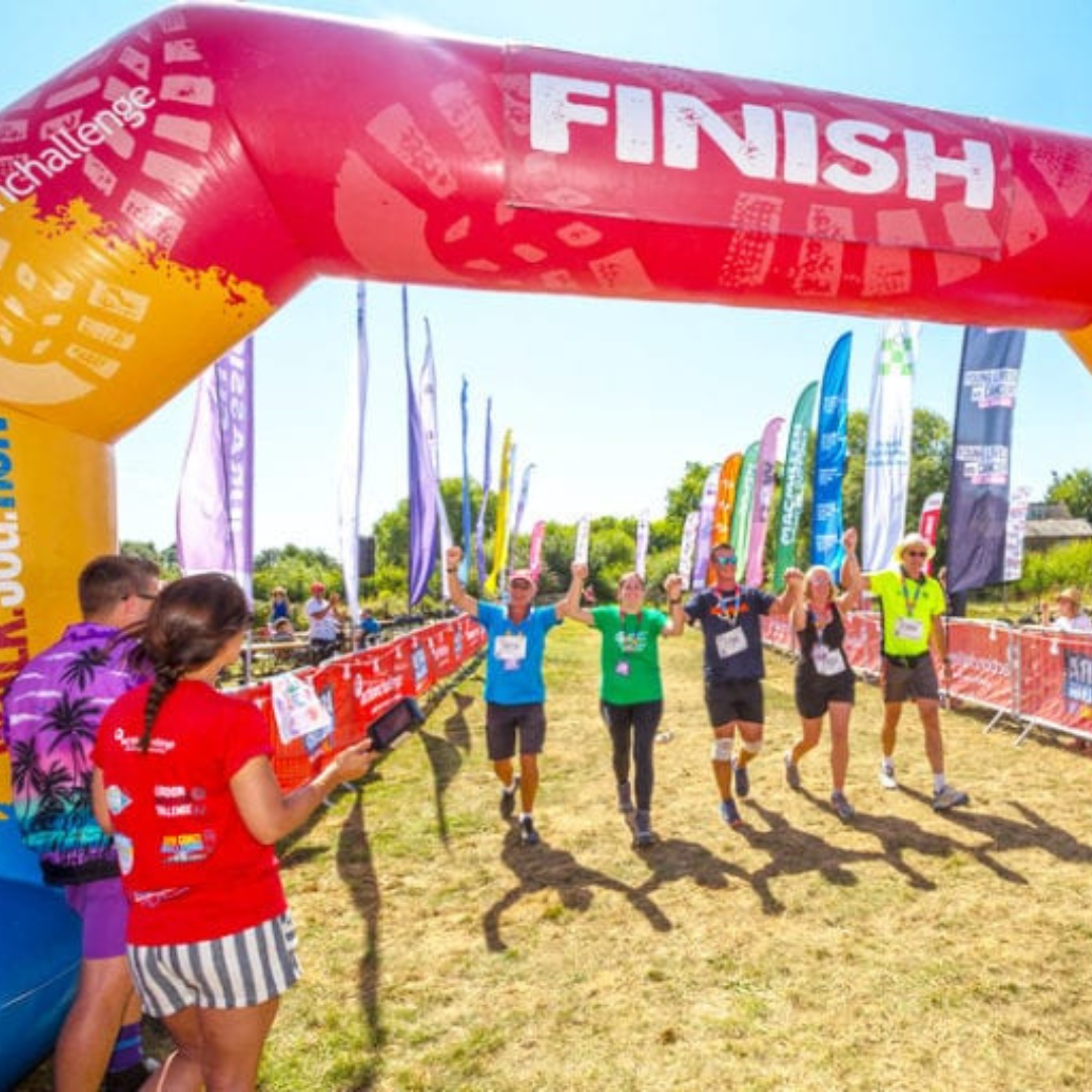 Runners cross a large red and yellow inflatable finish arch at an outdoor race under a clear blue sky, with spectators and event banners in the background.