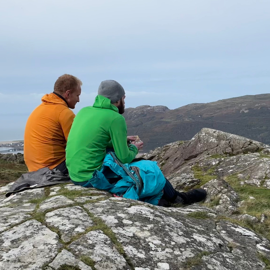Two people sitting on a rocky outcrop with a scenic view of mountains and water.