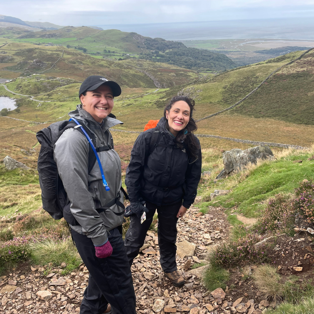 Two hikers on a trail with a scenic view of rolling hills and a lake.