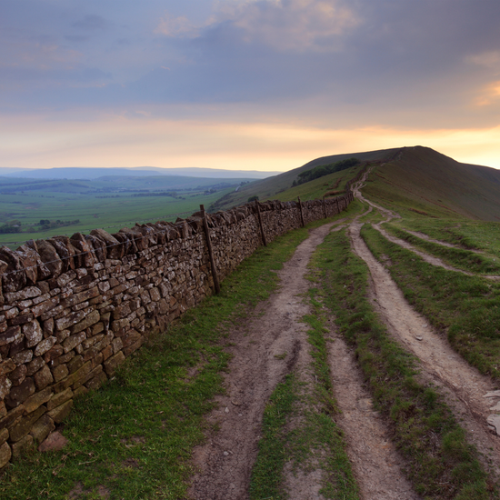 Winding dirt path along a stone wall with a scenic landscape and sunset in the background.