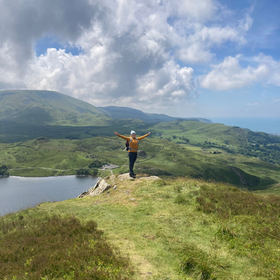 Person standing on a hill with arms outstretched, overlooking a scenic landscape with a lake and mountains.