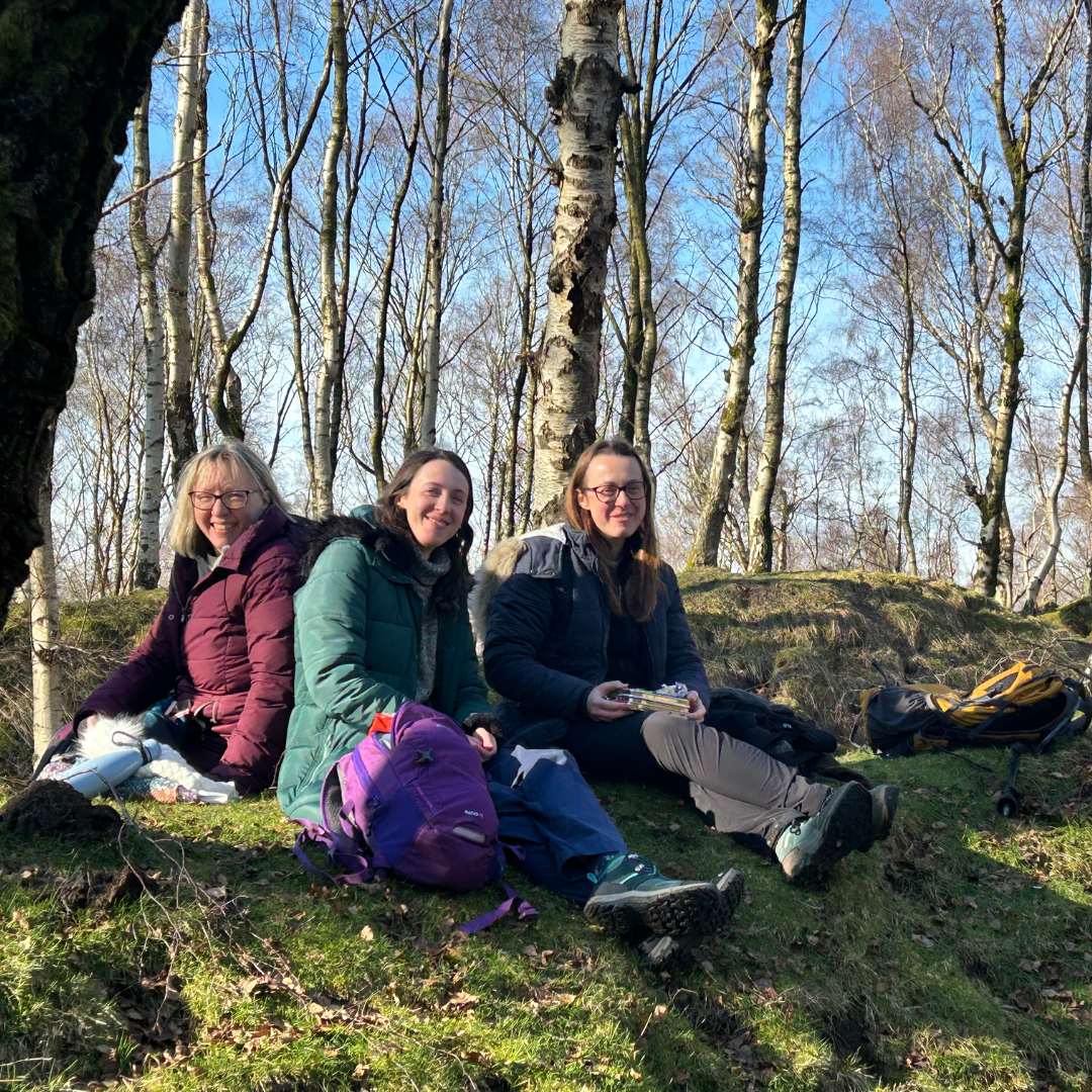 Three people sitting on a grassy hill with trees in the background