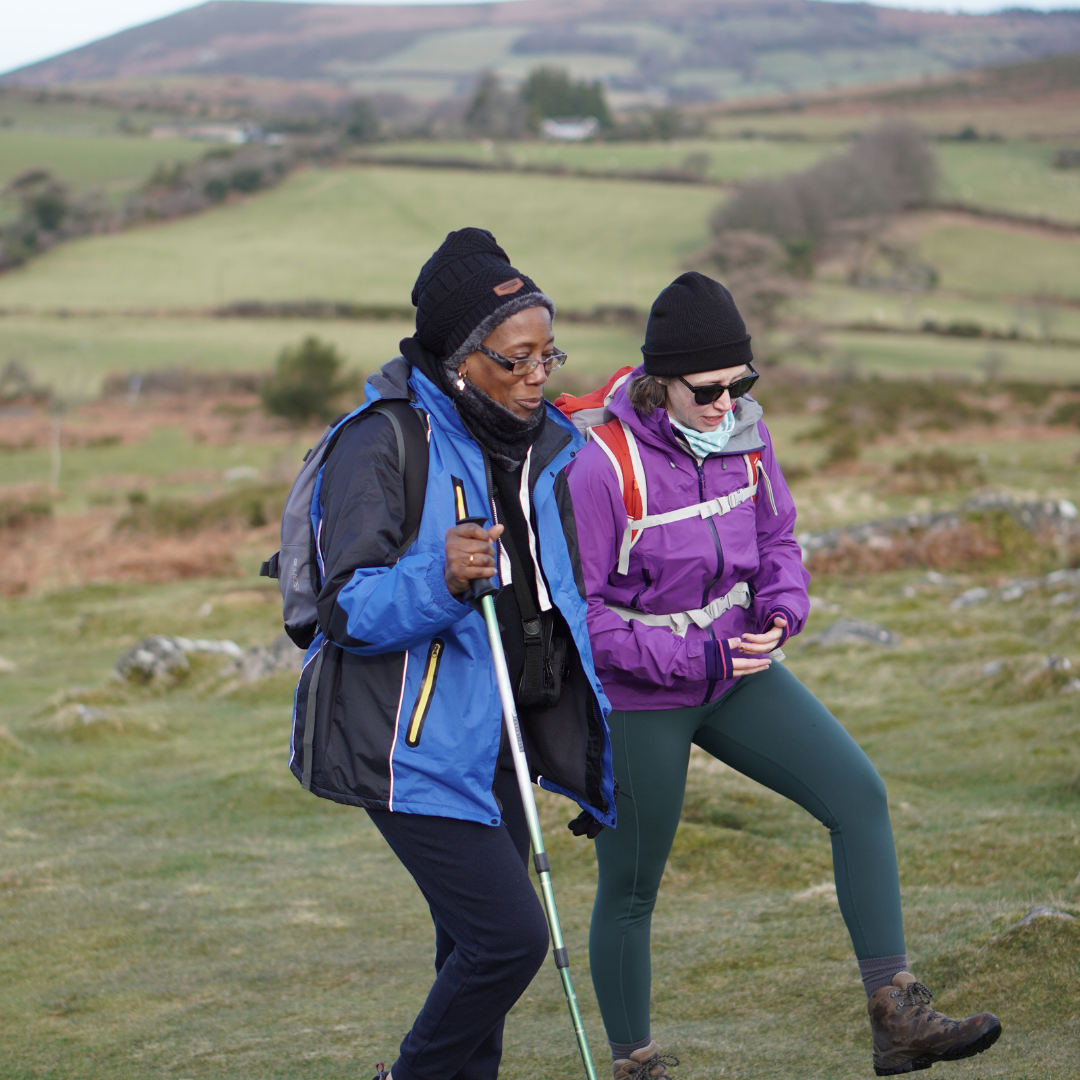 Two people hiking in a scenic outdoor setting with green fields and hills.