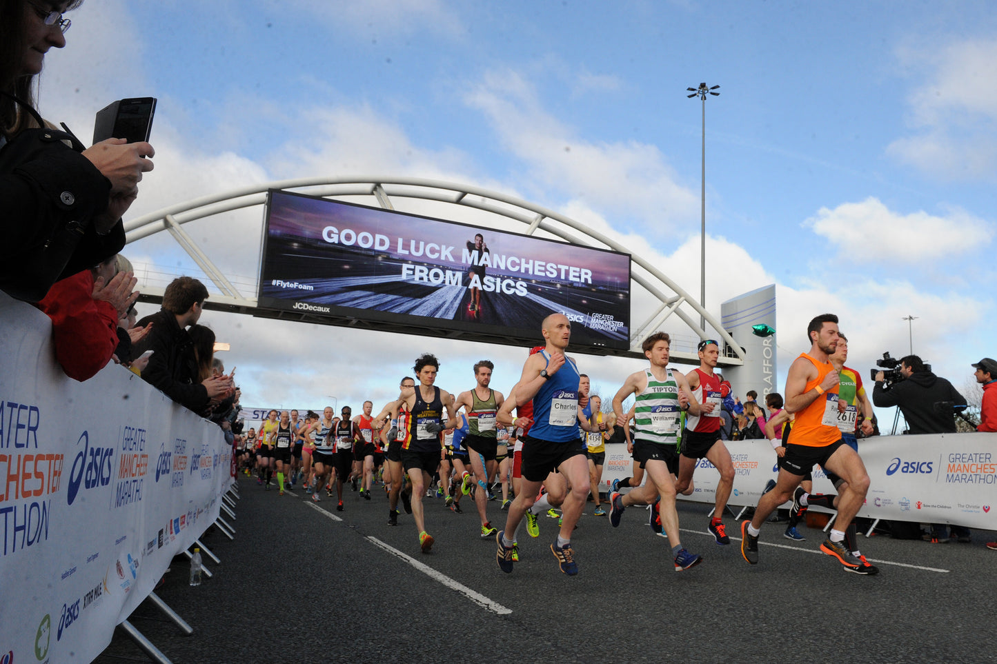 A group of marathon runners crosses the finish line under a large archway at the Manchester Marathon, with spectators lining the left side of the road. Visible branding includes ASICS on the barriers and finish line banner.