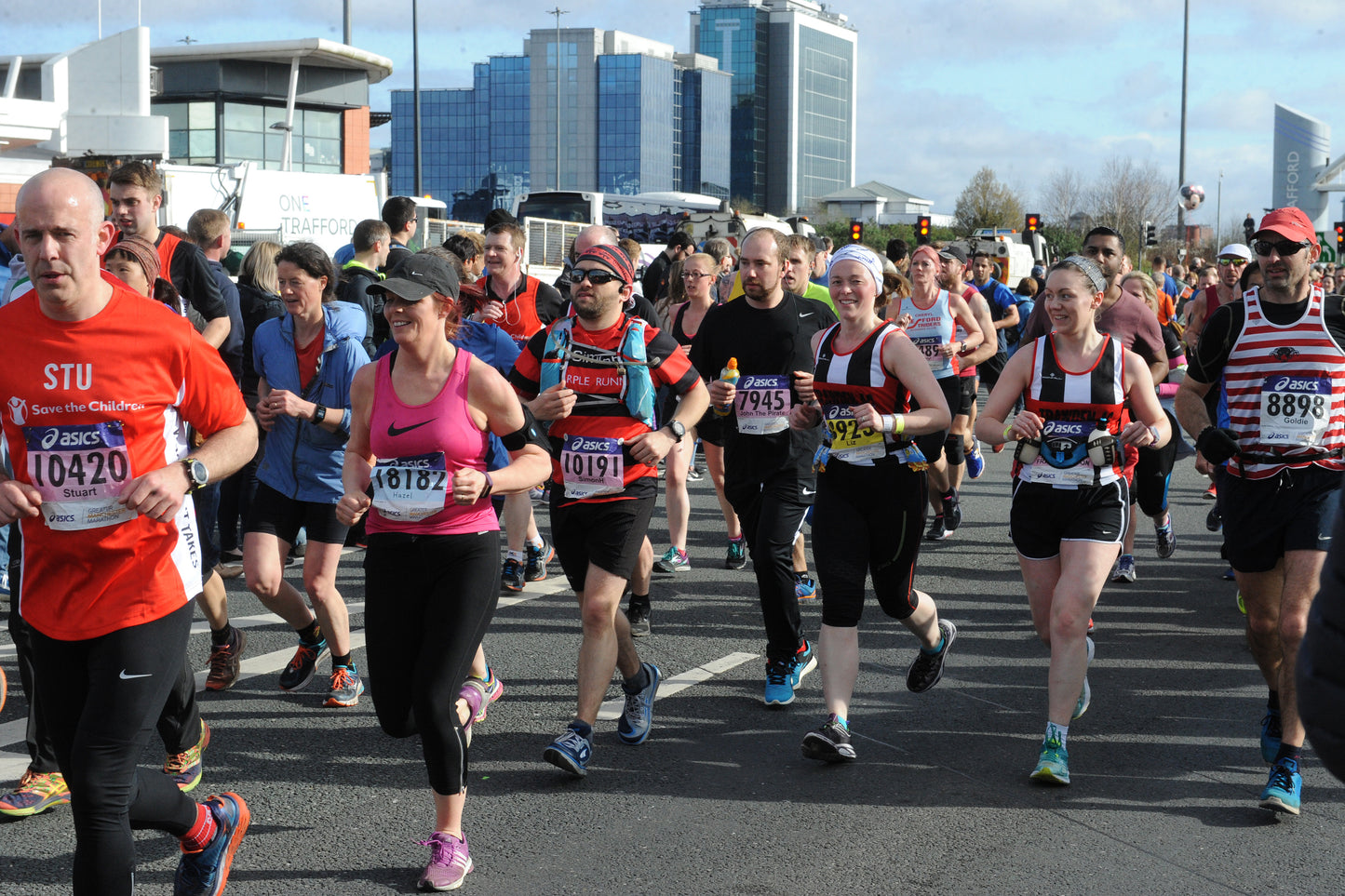 A group of marathon runners crossing a city street, with modern buildings in the background. Several runners are wearing athletic bibs, and the scene is captured in daylight.