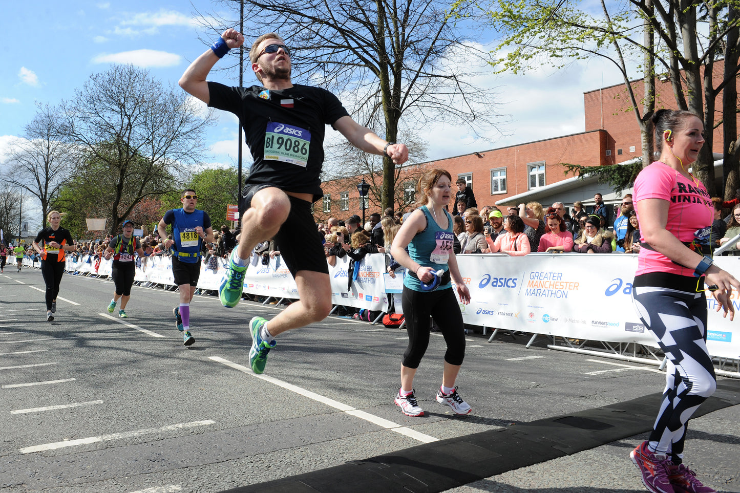 A man in a black t-shirt and black shorts jumps over a finish line tape at a road race, wearing a bib number 19066. Other runners, spectators, and trees are visible in the background.