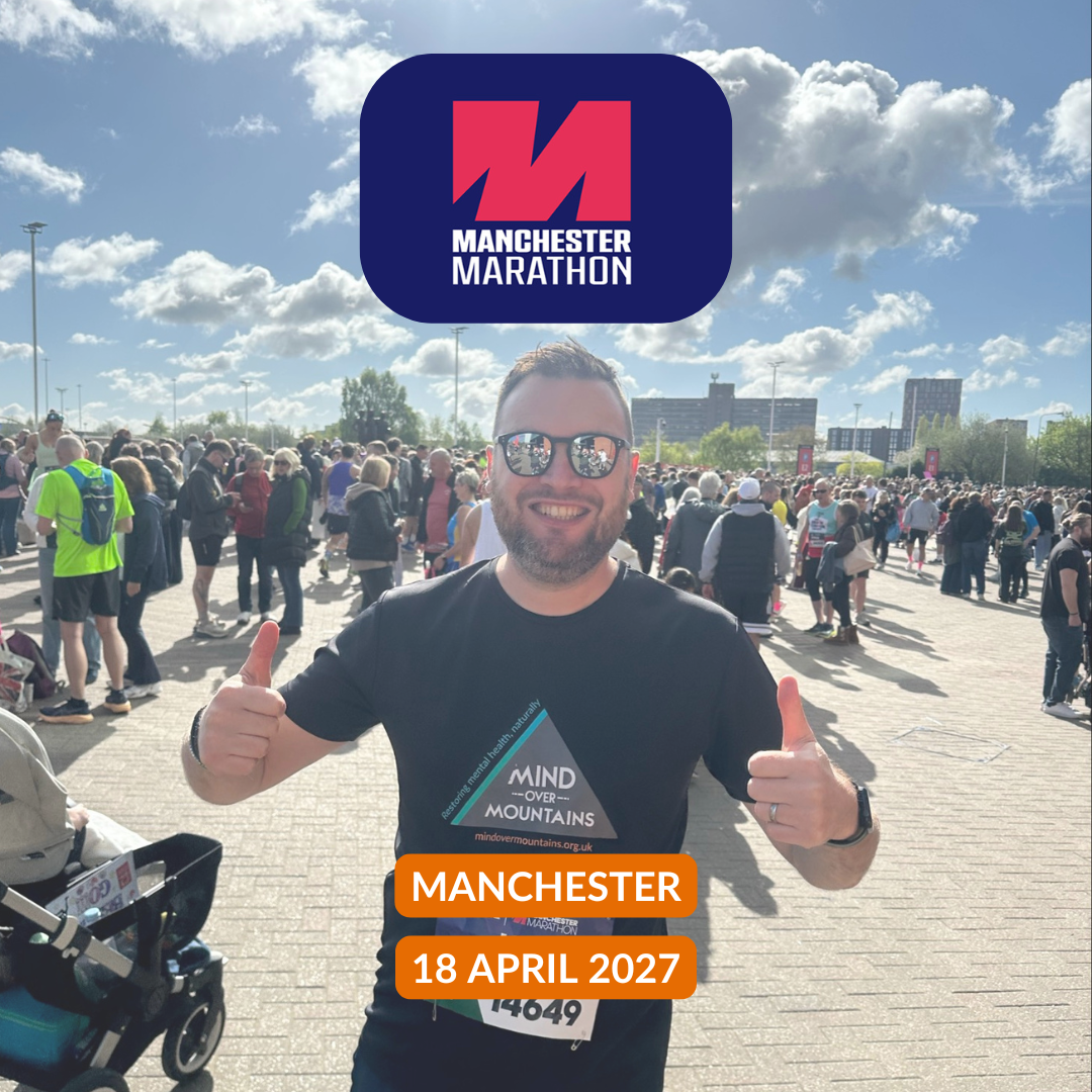 A man wearing a black Manchester Marathon t-shirt with a Mind the Mountains logo, giving two thumbs up in an outdoor crowd scene under a blue sky. A large Manchester Marathon logo is overlaid at the top, and an orange badge at the bottom reads Manchester Marathon 18 April 2027.