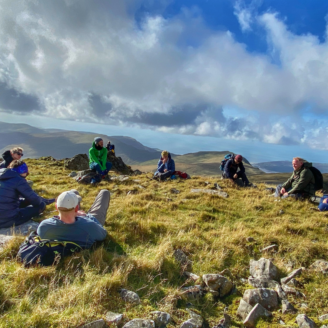mindfulness in nature in mountains - group of hikers relaxing in grass at top of mountain with mindfulness coach